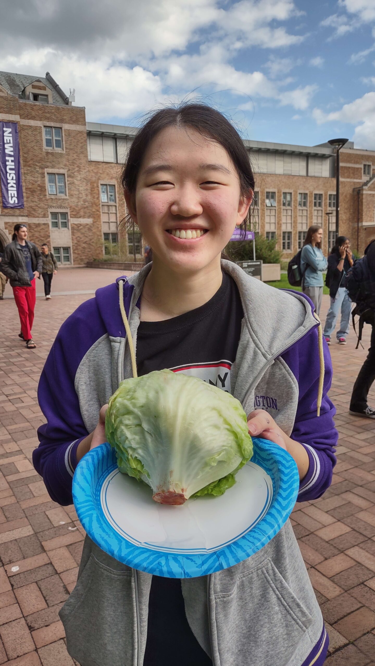 Smiling person holding a plate with a lettuce head, University of Washington campus in background